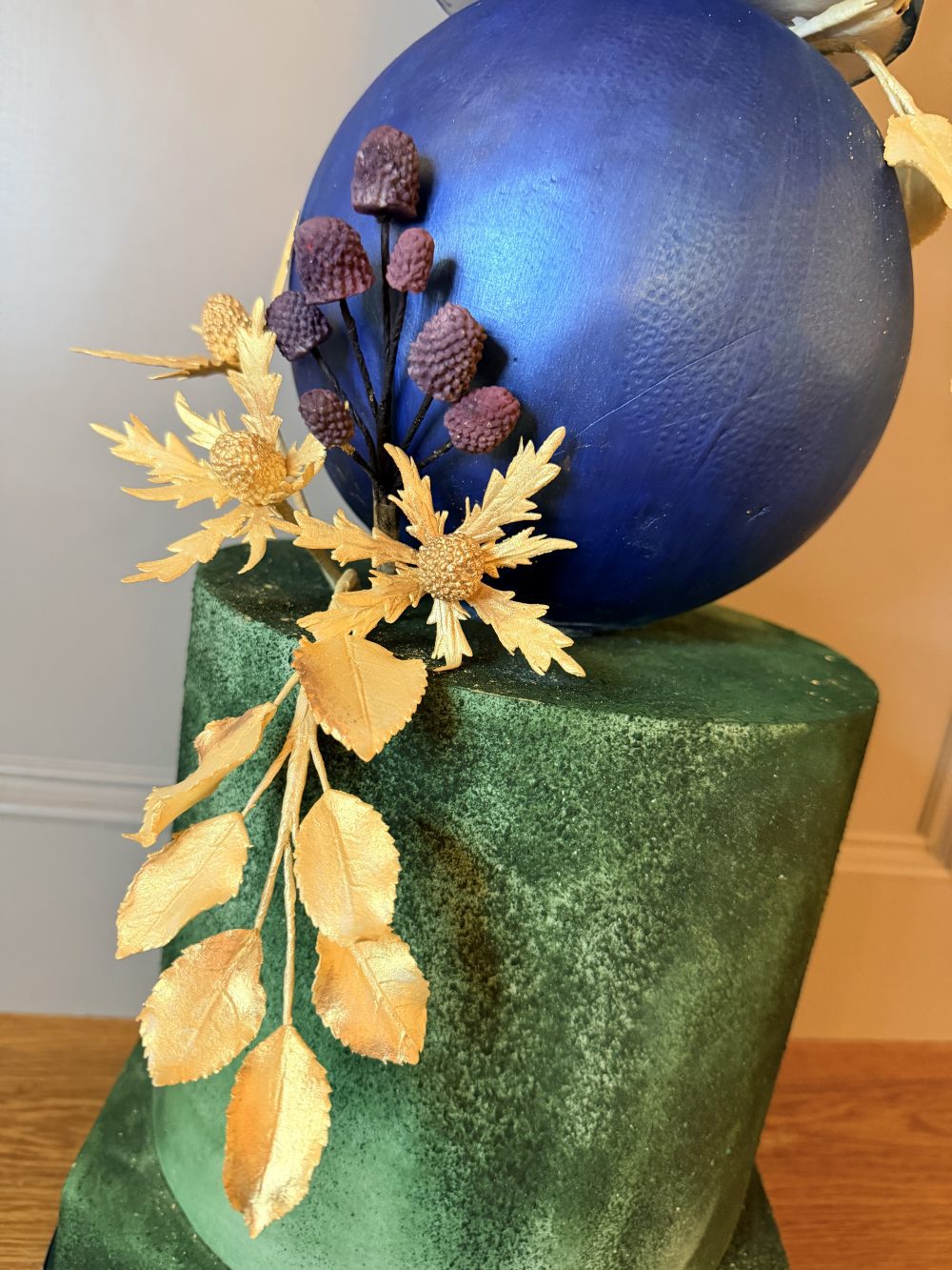 Close up detail of a modern wedding cake showing textured forest green velvet frosting contrasted against a metallic cobalt blue sculptural sphere with gold leaf accents.