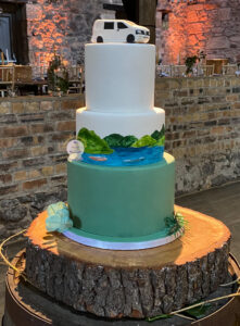 Bespoke wedding cake with a handmade camper van topper and painted landscape featuring the couple’s paddleboards.