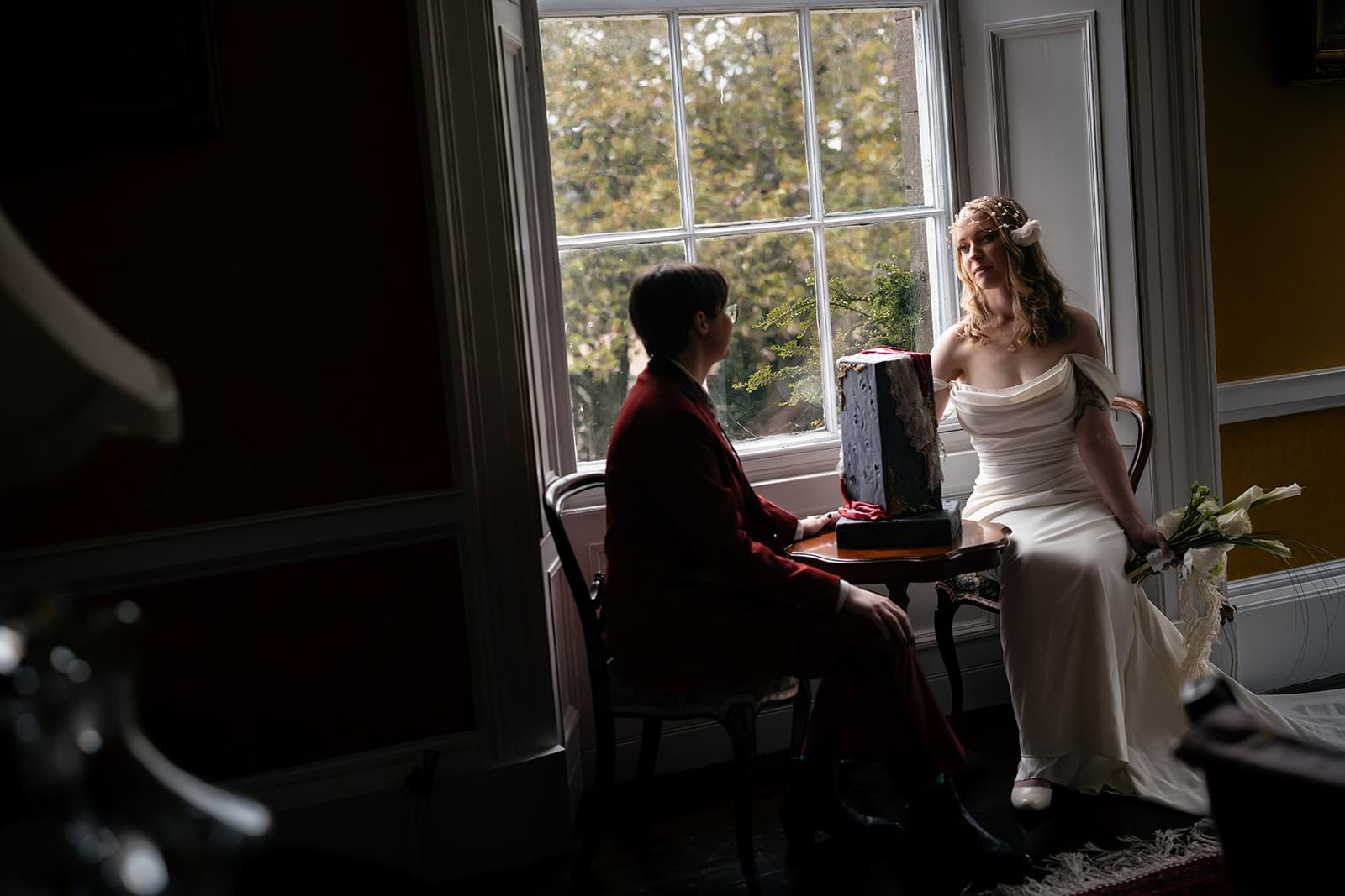 Bespoke luxury wedding cake by Crumbdidilyumptious, set against a light-filled grand window at a historic Perthshire estate with the couple seated nearby. Photography by The Snashfolds